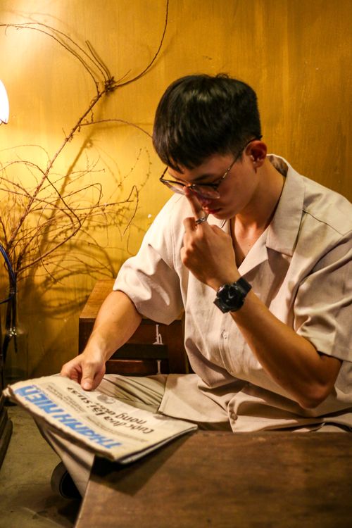 Free A young man in glasses concentrates on reading a newspaper, seated indoors in warm lighting. Stock Photo