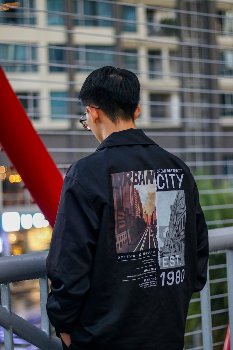 Young Man In A Jacket With Graphic On The Back Standing In City 