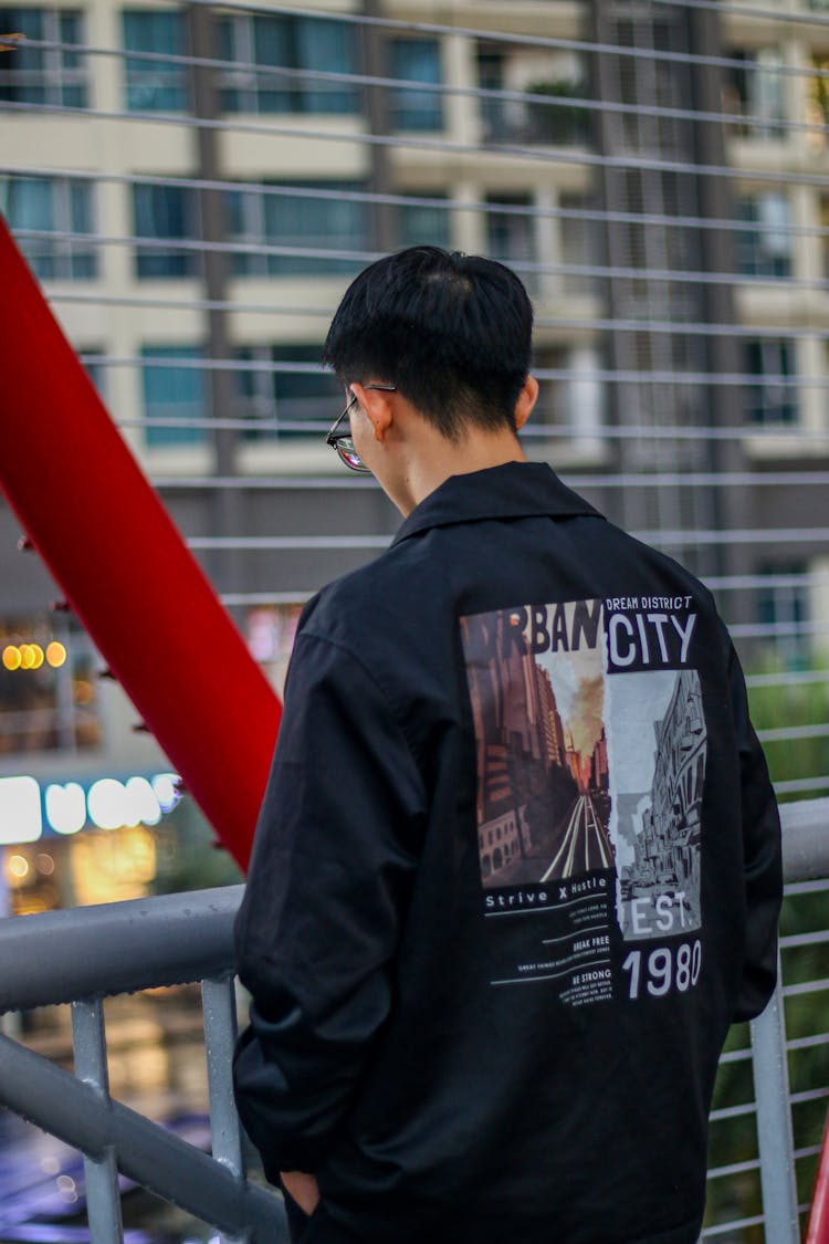 Young Man In A Jacket With Graphic On The Back Standing In City 