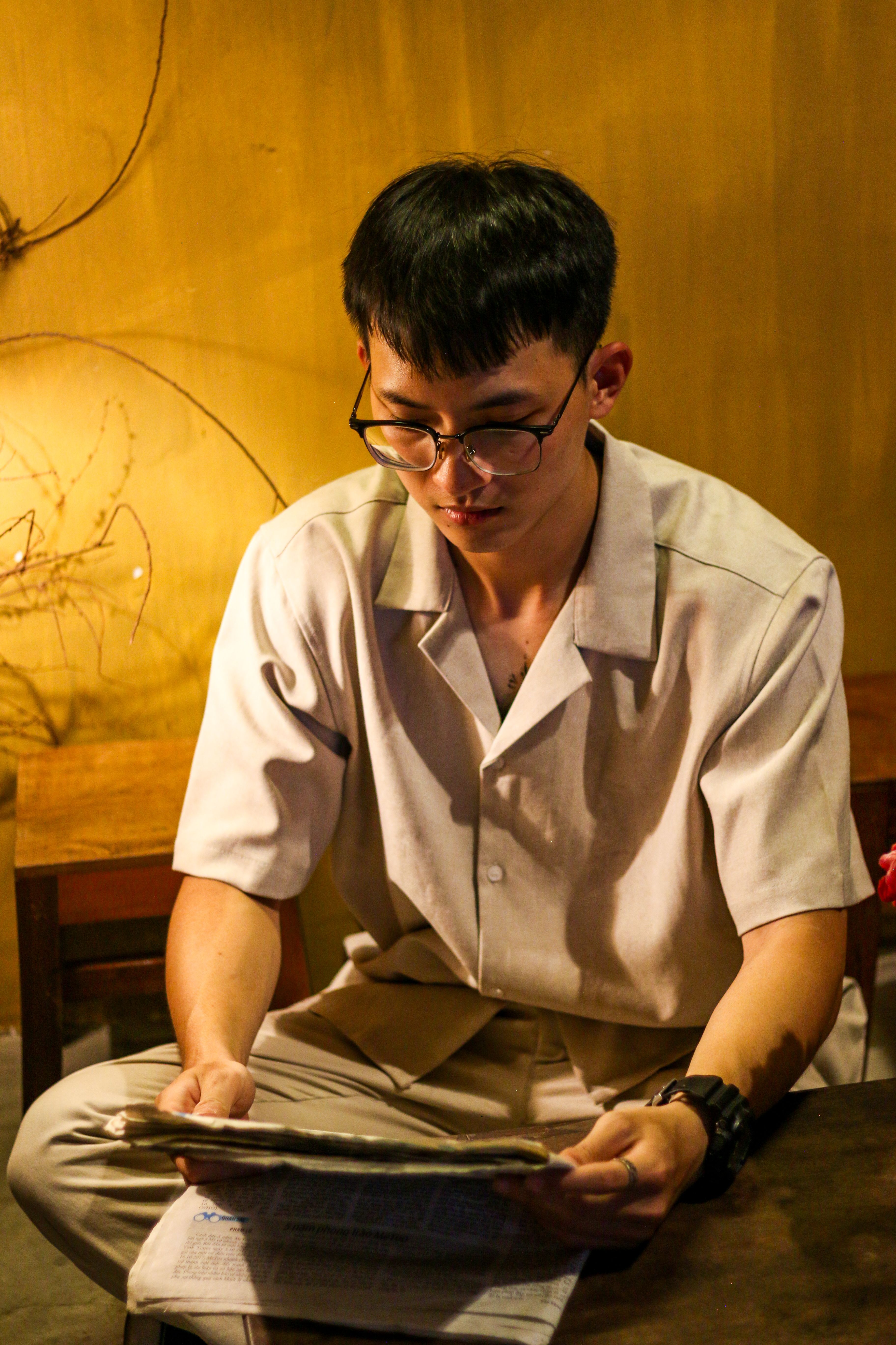Asian man with eyeglasses reads newspaper in a warmly lit room.