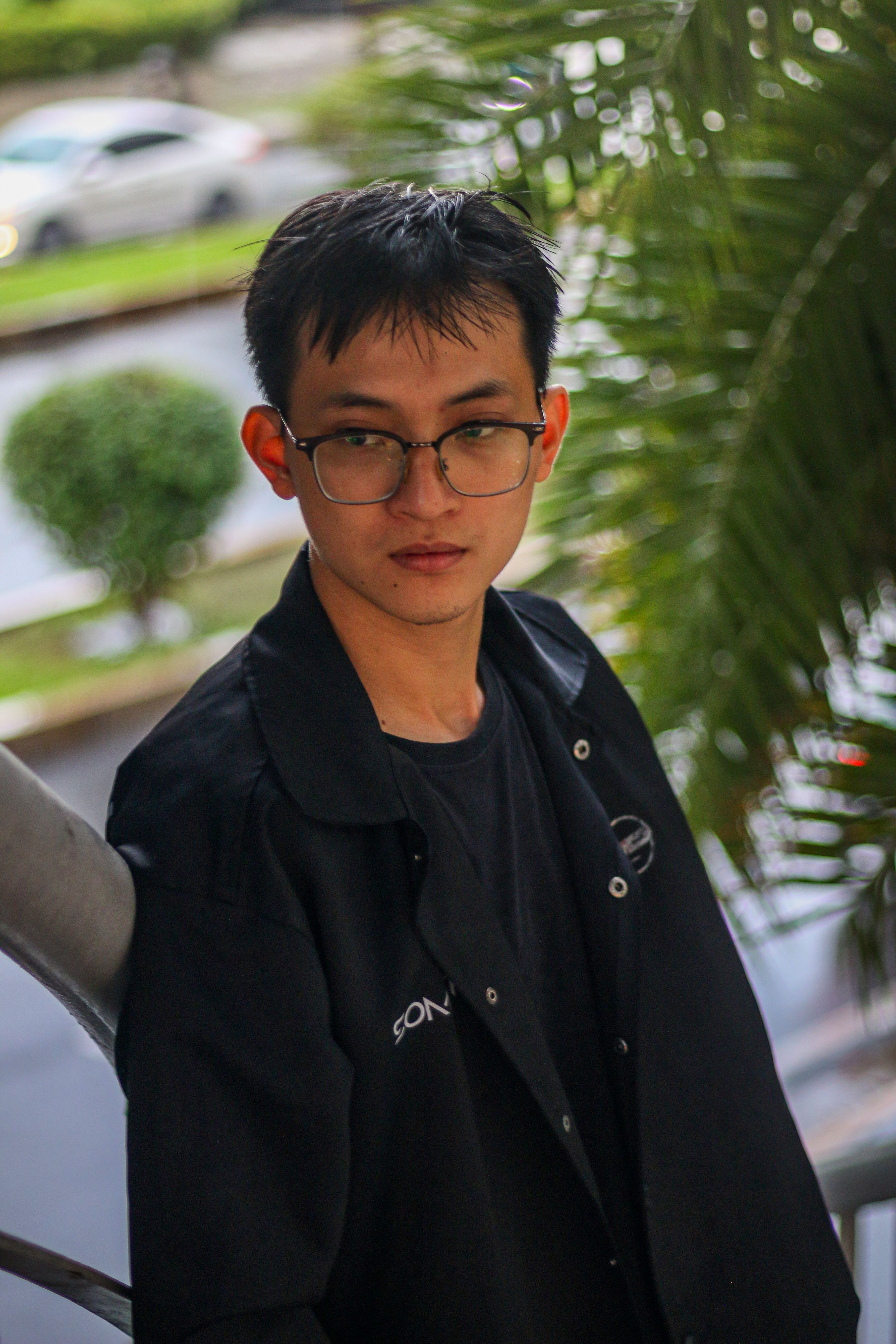 Portrait of a young man with glasses in a black jacket, standing outdoors with urban elements and greenery.