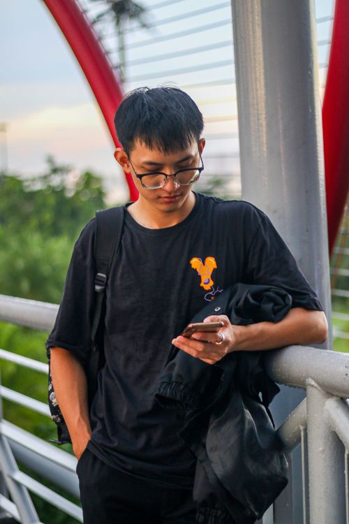Free A young man standing on a bridge, engaged with his smartphone, set against a vibrant urban backdrop. Stock Photo