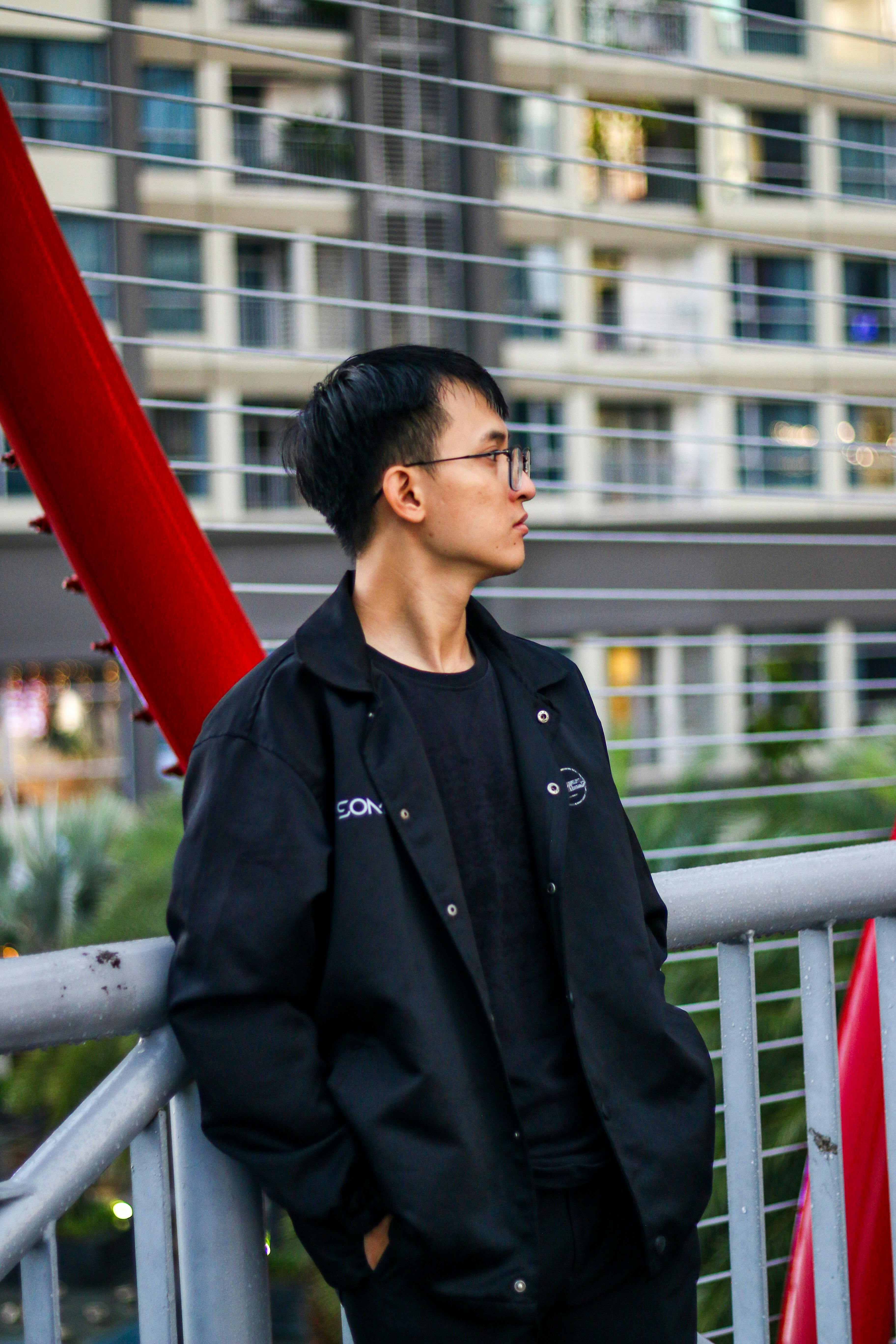 A young man in a black jacket stands on an urban balcony, gazing into the city view.