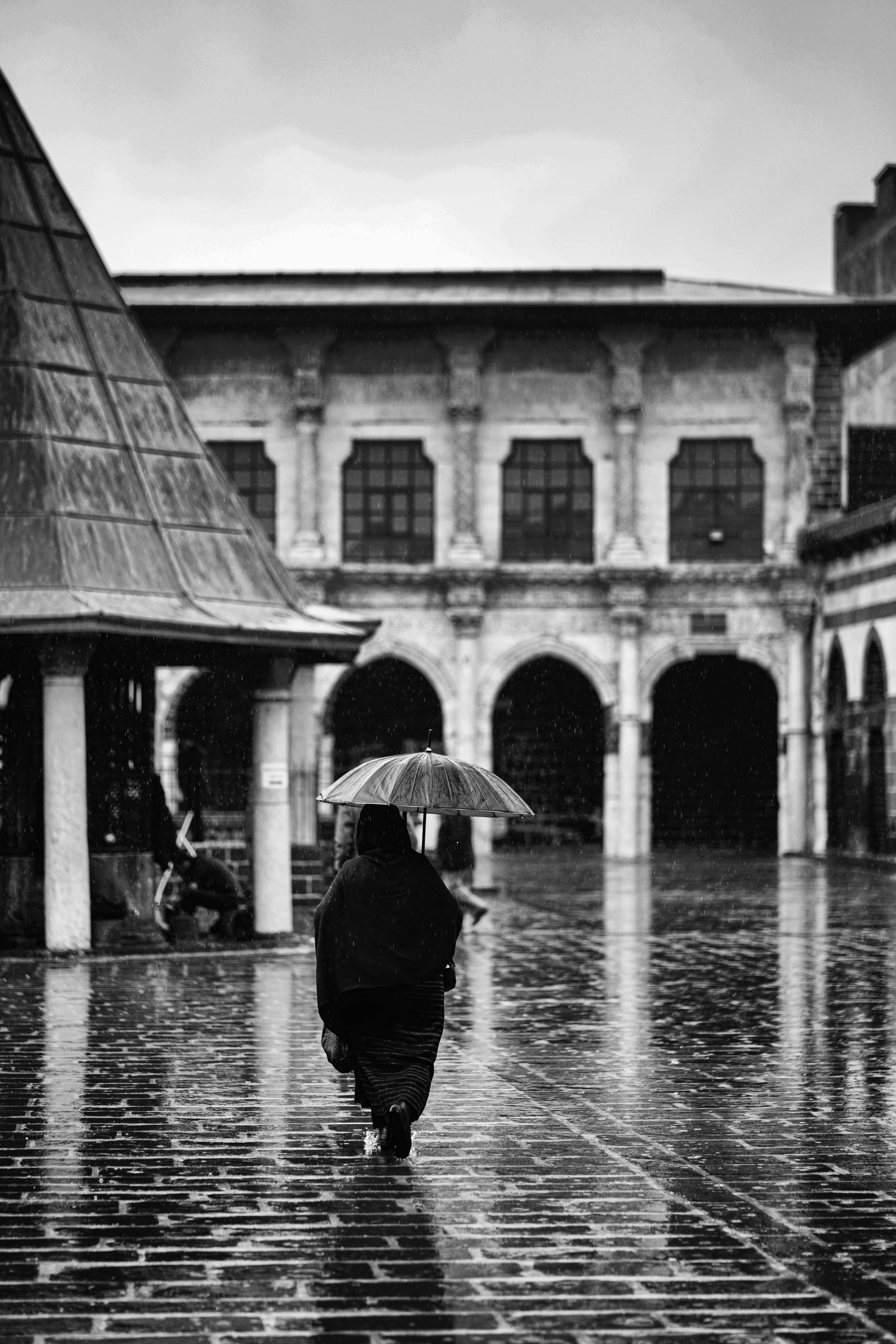 Black and white image of a woman with an umbrella walking in a historical courtyard on a rainy day.