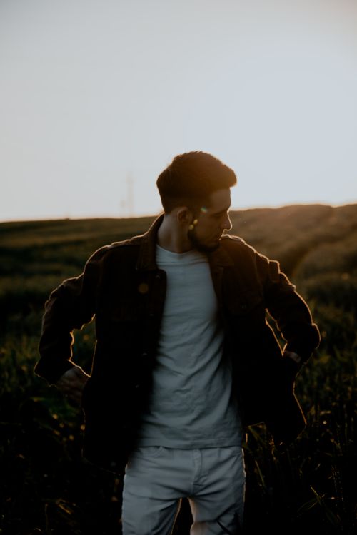 Free Silhouette of a young man standing in a field at sunset, creating a dramatic and moody atmosphere. Stock Photo
