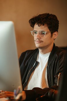 Focused man with eyeglasses and jacket working on computer indoors.