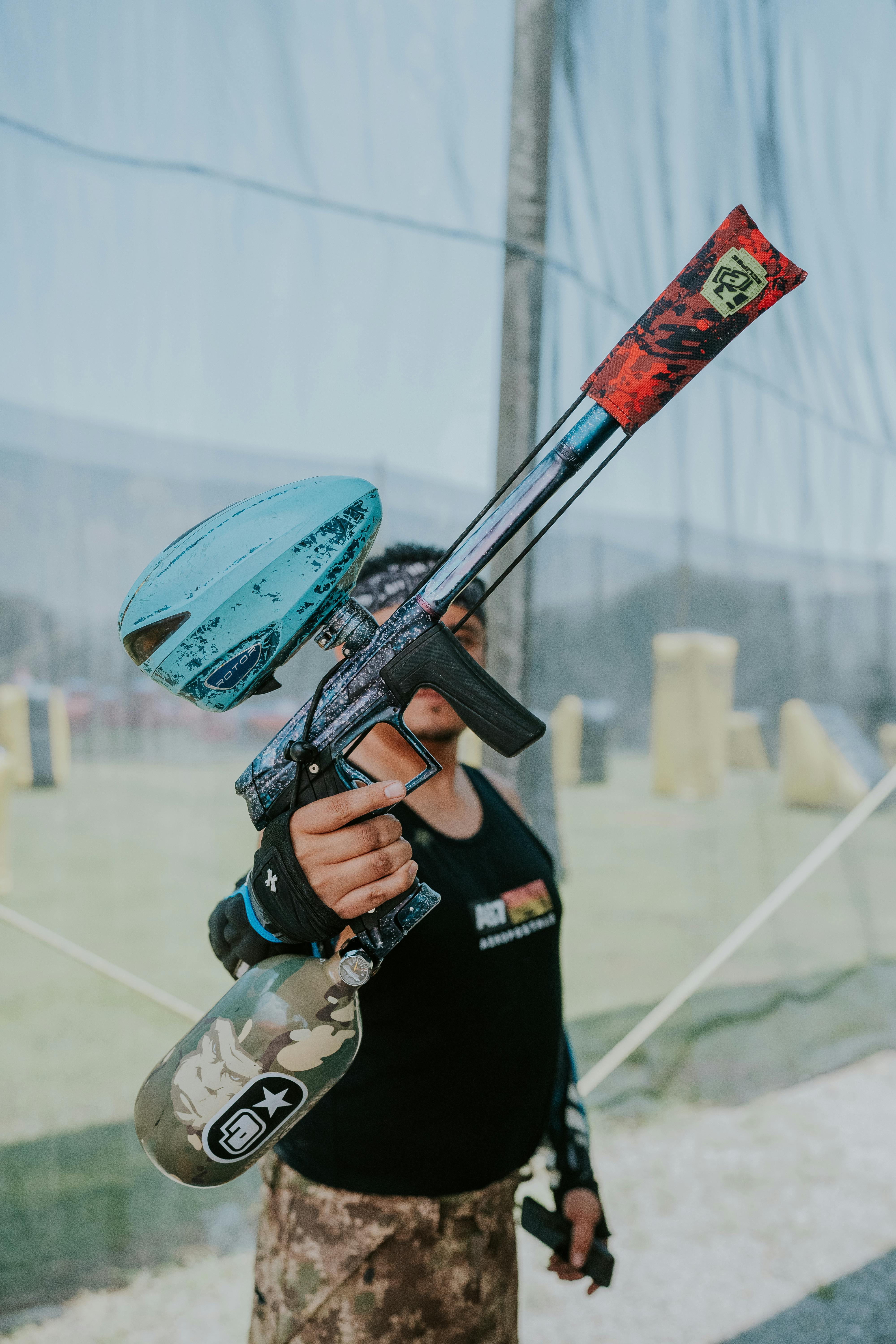 Un homme se tient dehors pendant la journée, tenant un pistolet de paintball coloré. Prescription Goggle Inserts - A man stands outdoors during the day, holding a colorful paintball gun.