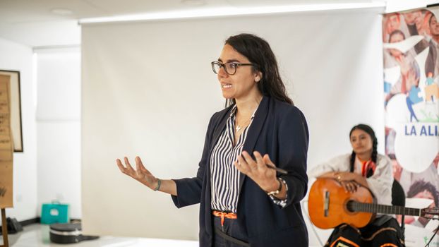 Woman presenting at an indoor business event with a guitar player in the background.