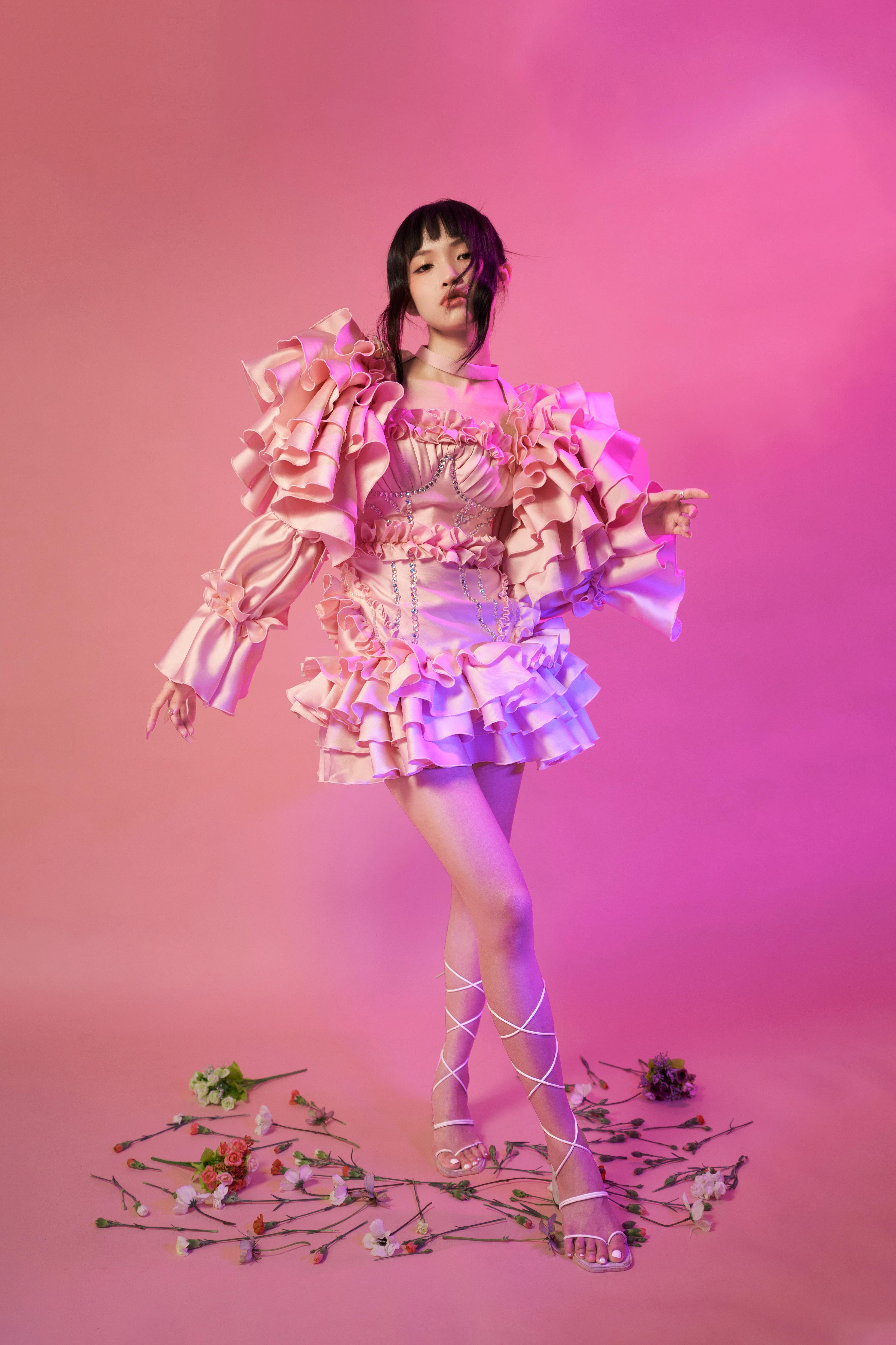 A stylish fashion portrait of a woman in a ruffled pink dress with floral elements and a pink background.