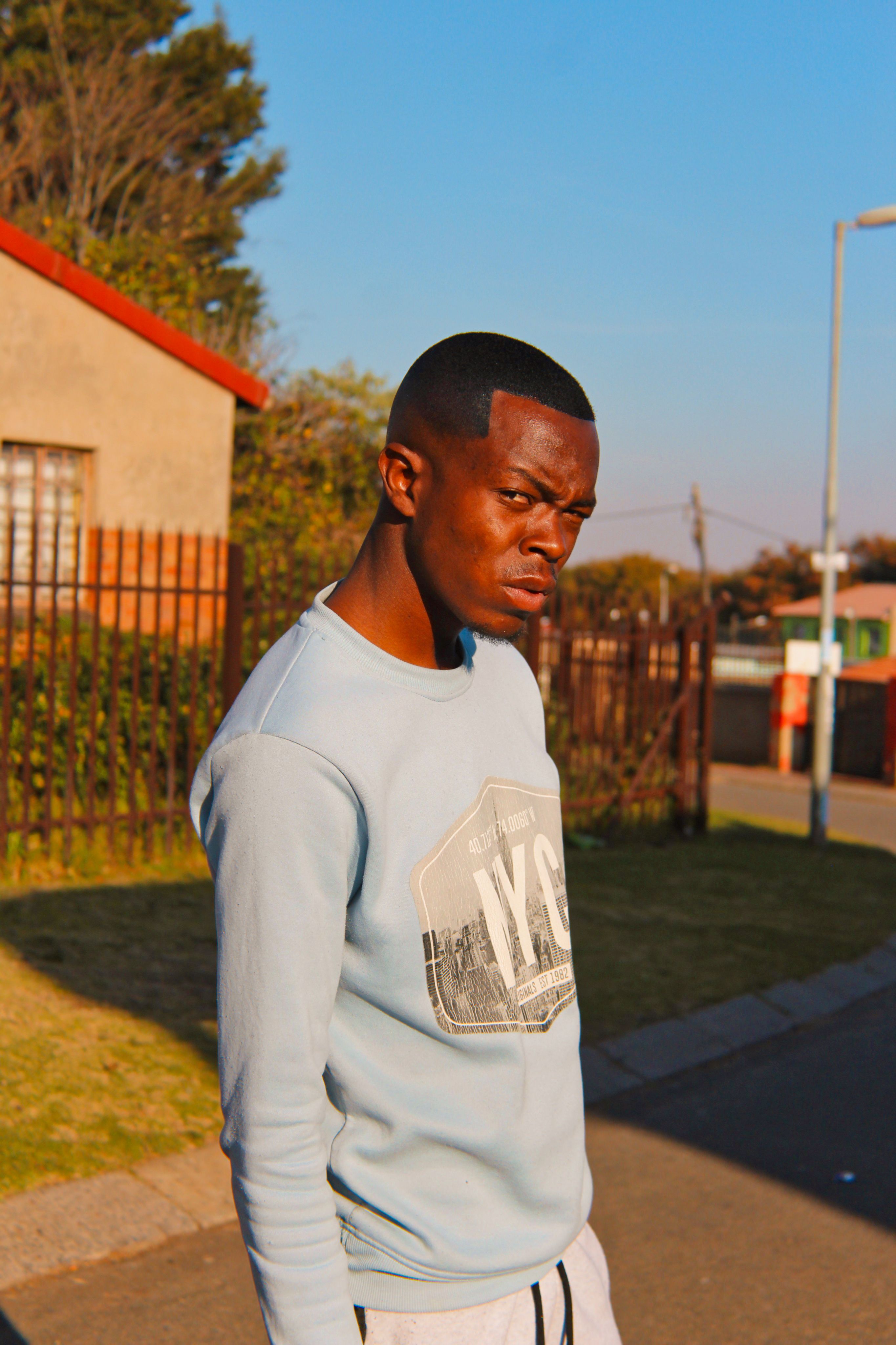 A man with short hair stands on a sunlit urban street, exuding confidence.