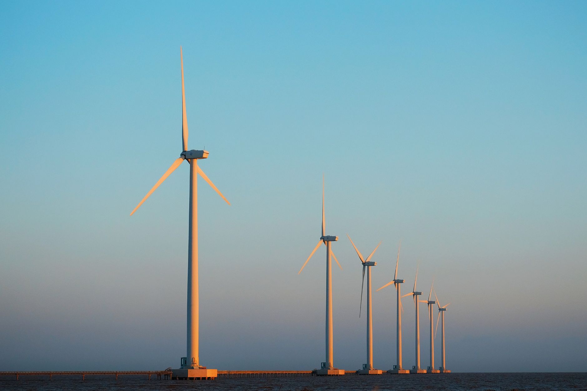 A wind farm with several turbines lit by the sunset by the sea, symbolizing renewable energy.