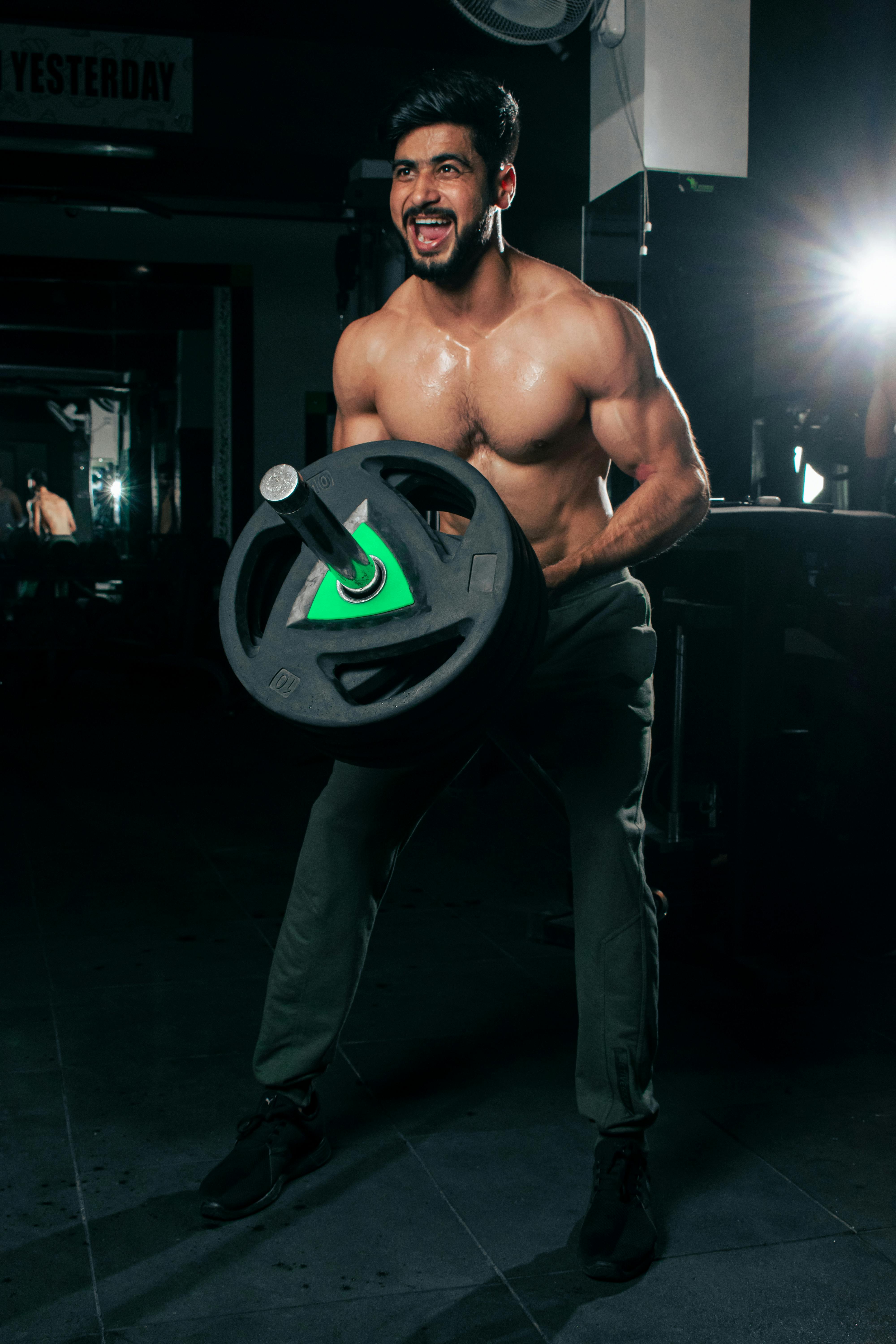 A muscled man performs weight lifting exercises in a dimly lit gym environment, showcasing strength and focus.