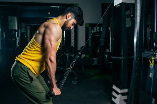 A fit man in a yellow tank top exercises using a cable machine in a gym setting.