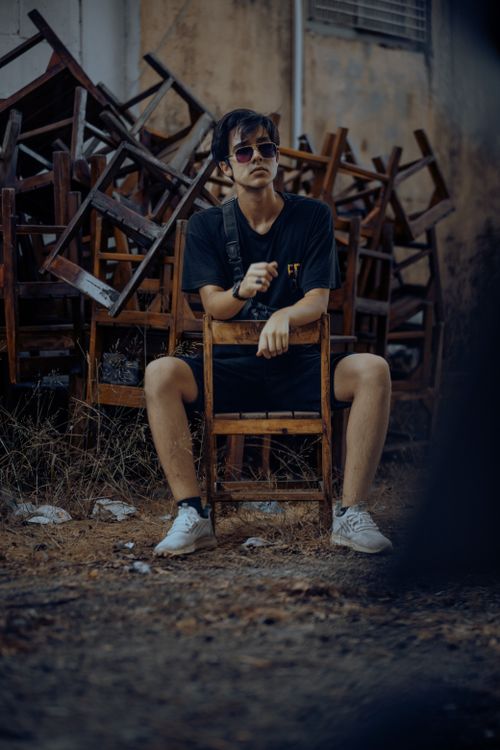 Free Stylish young man in sunglasses sits confidently amid stacked wooden chairs. Stock Photo