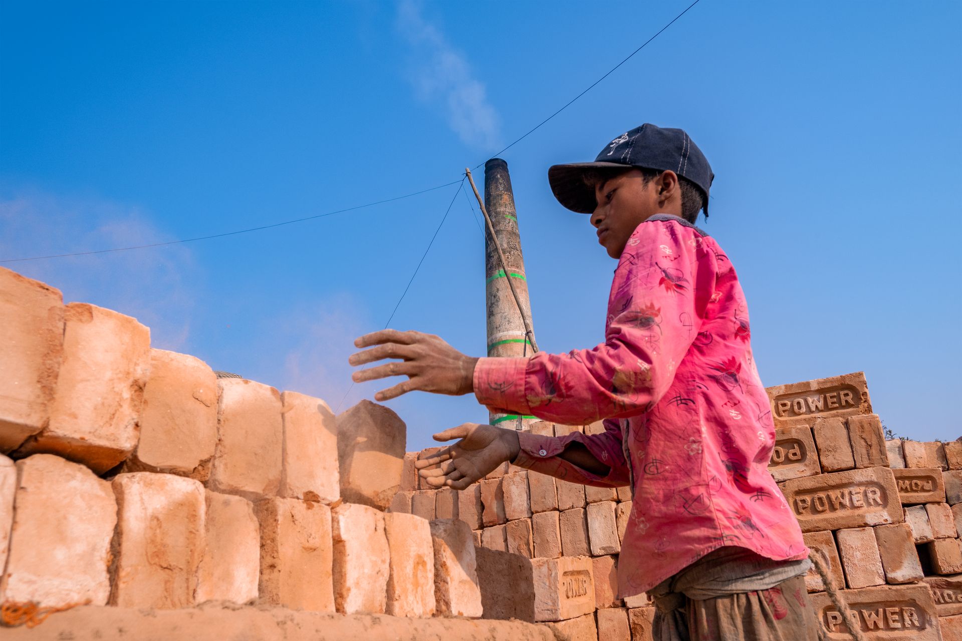 A child wearing a pink shirt and cap working in an outdoor brick factory in India.