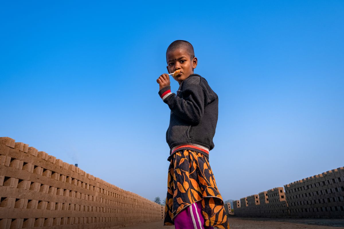 A young boy stands outdoors enjoying a lollipop against a bright blue sky. Urban setting.
