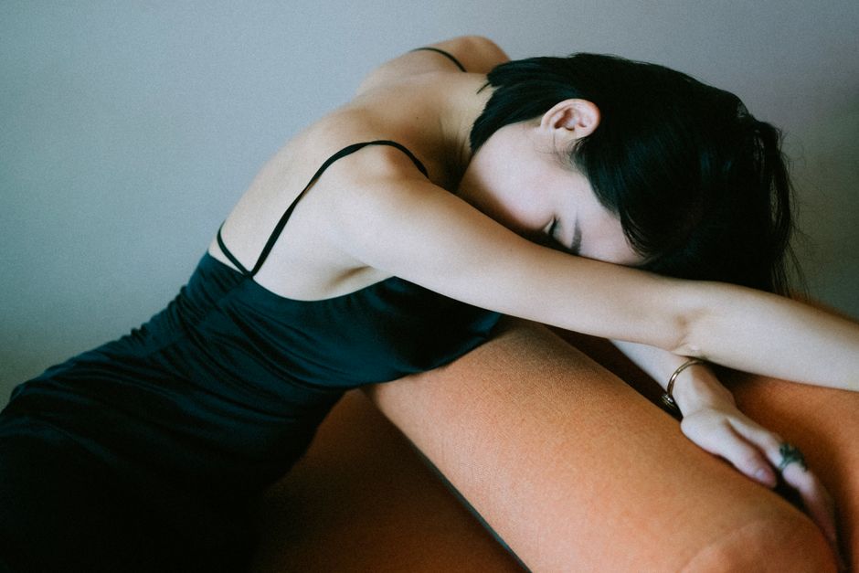 A woman in a black dress leans forward on a couch armrest, creating a contemplative and serene mood.