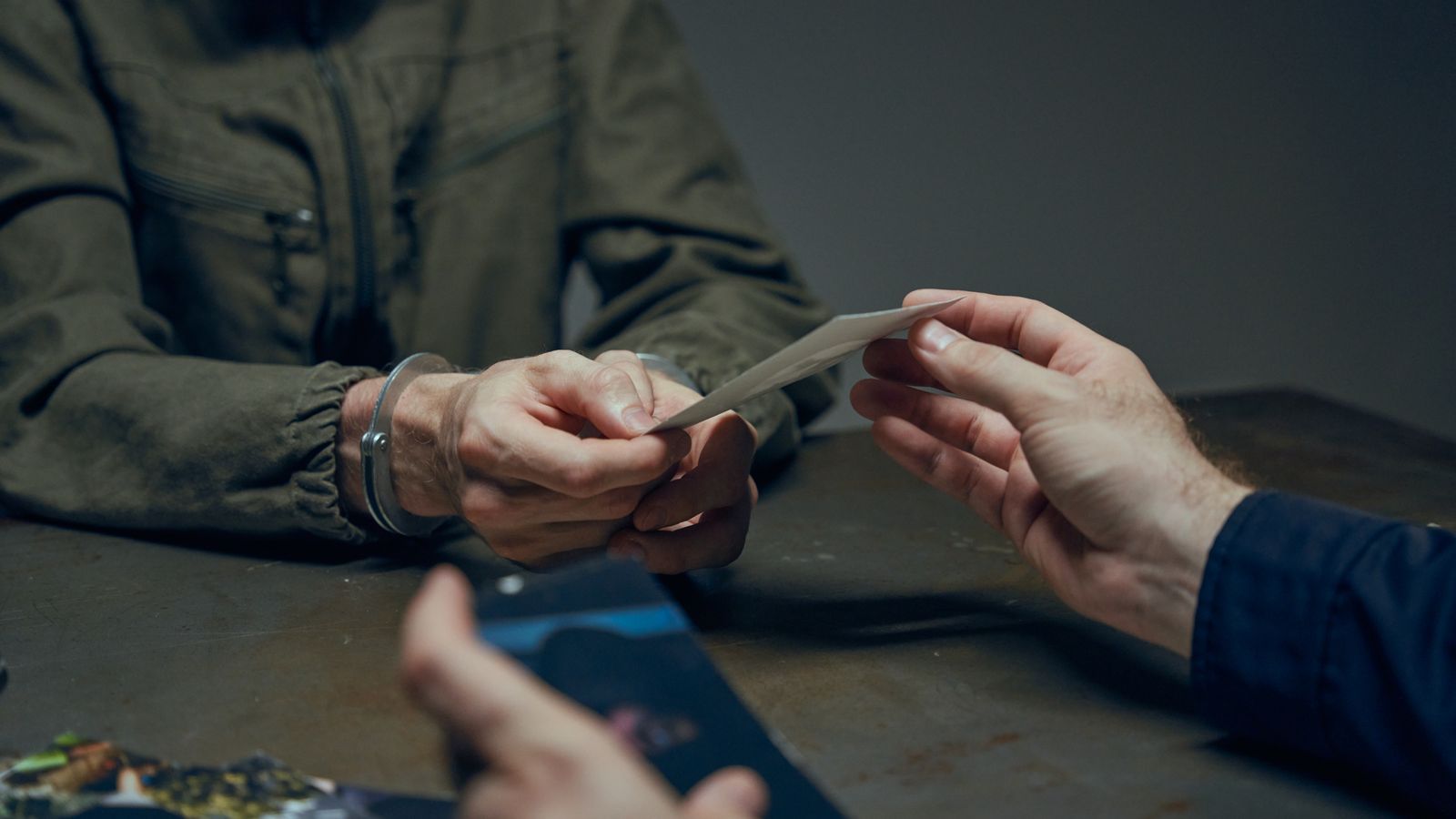 Hands exchanging evidence during an interrogation scene in a dimly lit room.