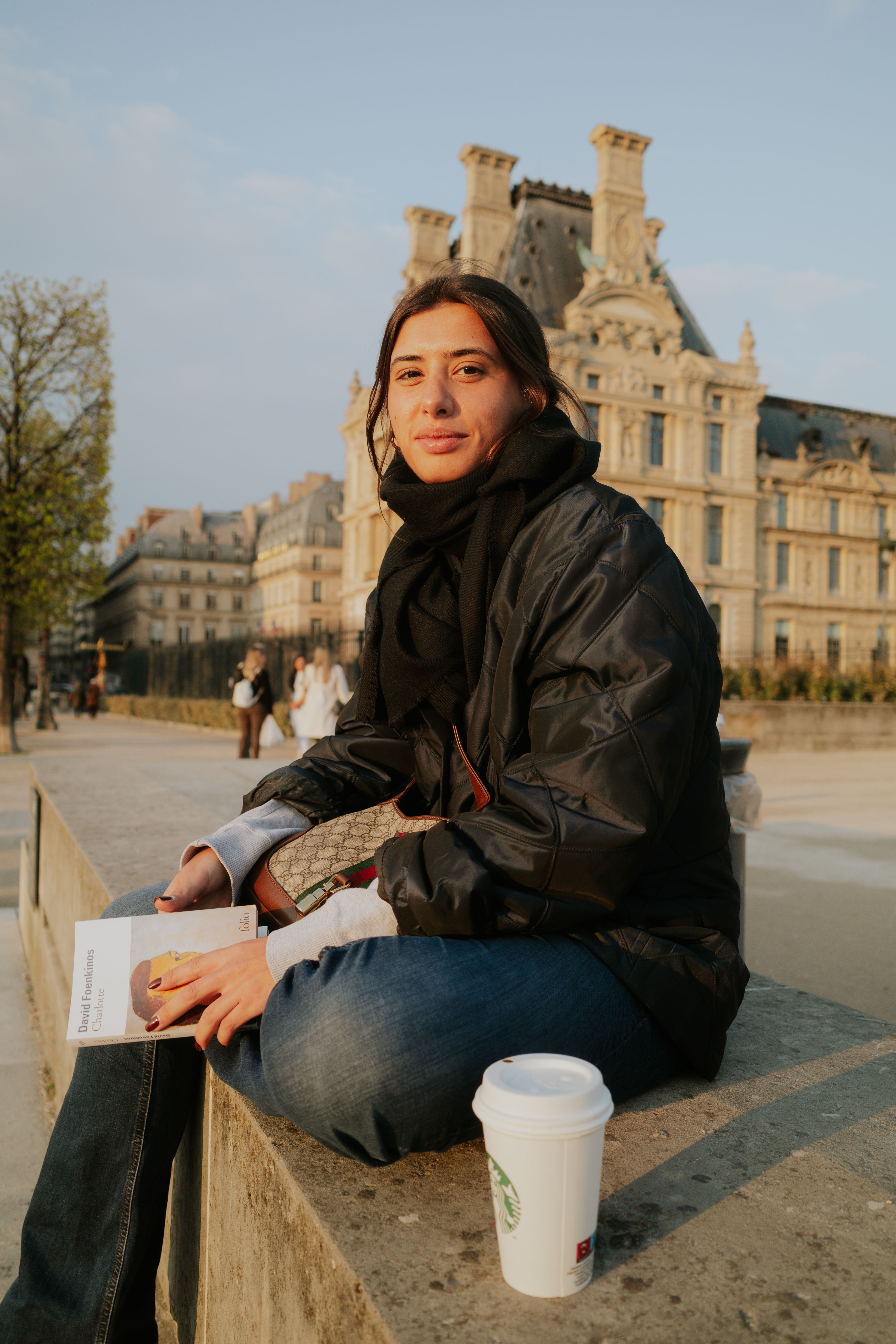 A woman in a black jacket enjoys a coffee outside a historic building.