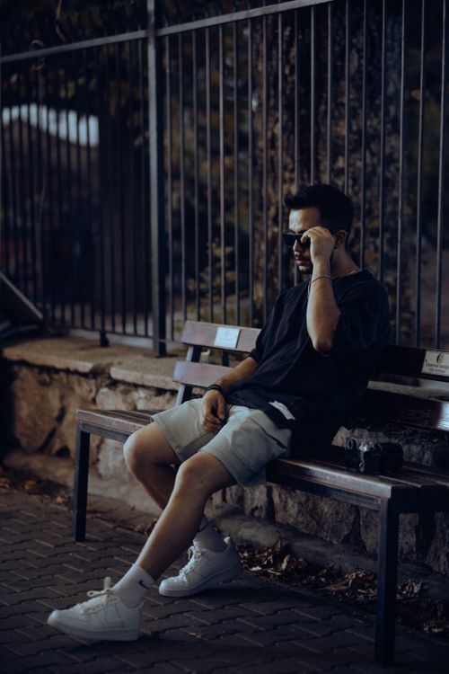 Free A young man in sunglasses and casual attire poses on a city bench at twilight. Stock Photo