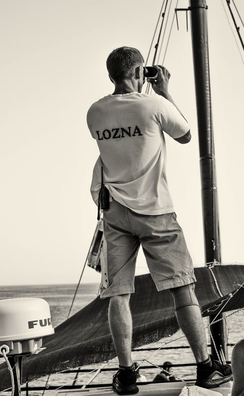 Free A sailor stands on a sailboat deck, using binoculars to scan the horizon, showcasing nautical adventure. Stock Photo