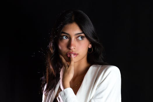 A young woman with long brunette hair wearing an elegant white blazer in a studio shoot with a black background.