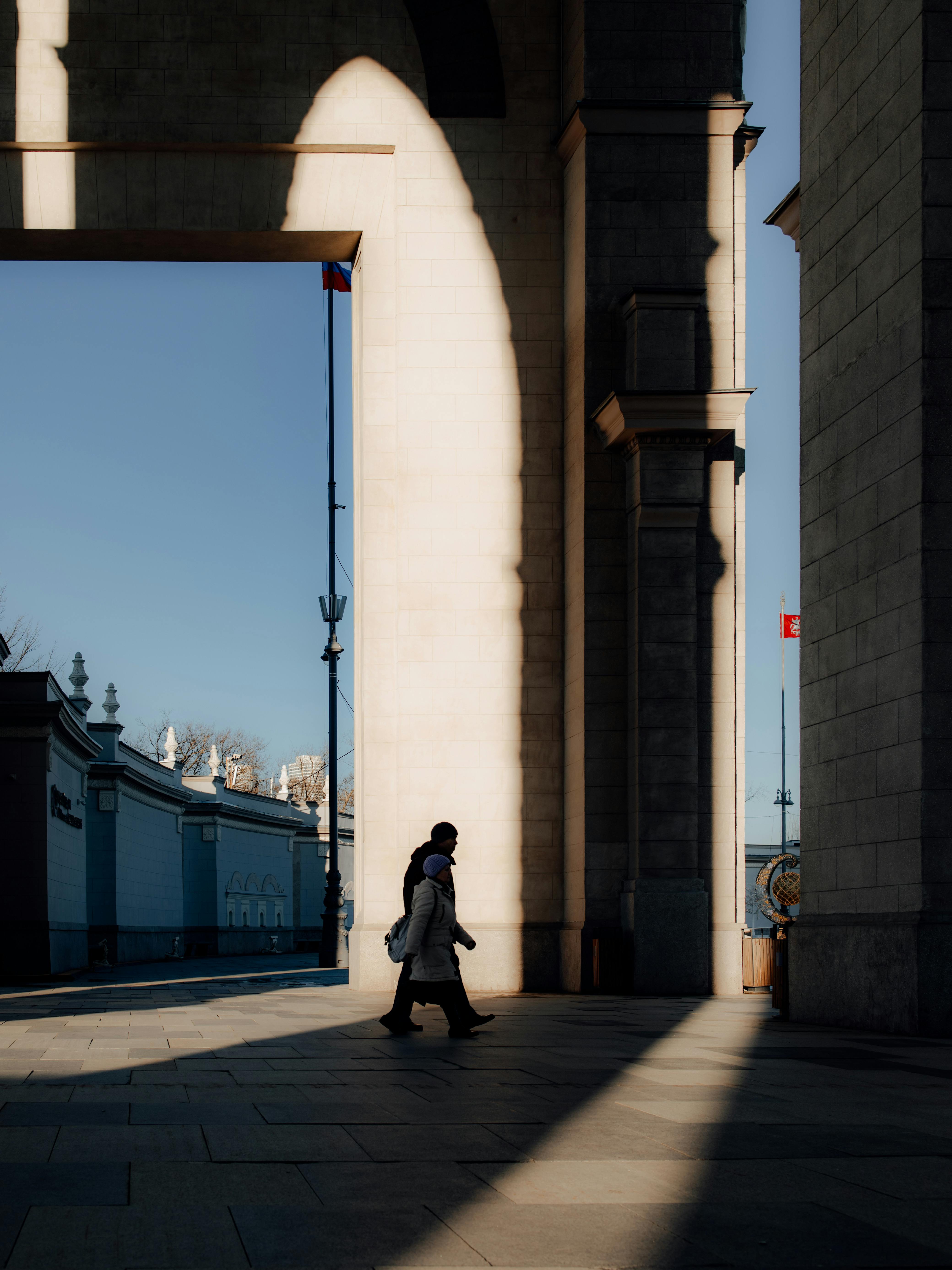 A lone figure walks through a sunlit archway, casting a dramatic shadow on a clear day.
