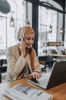 A woman in a hijab multitasks with a phone and laptop in a stylish café, showcasing modern working culture.