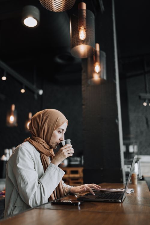 Free Woman in headscarf enjoys coffee while working on a laptop at a stylish café. Stock Photo