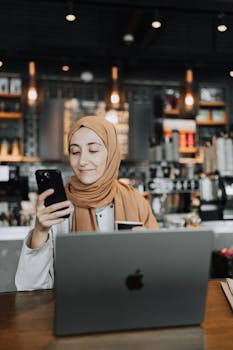 A woman in a hijab is smiling while using a smartphone at a modern café with her laptop.