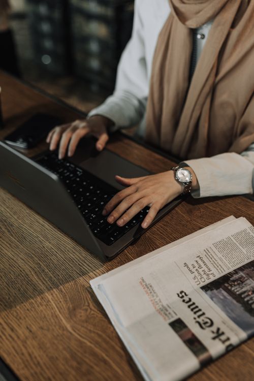 Free Woman working on a laptop at a wooden desk with a newspaper beside her. Stock Photo