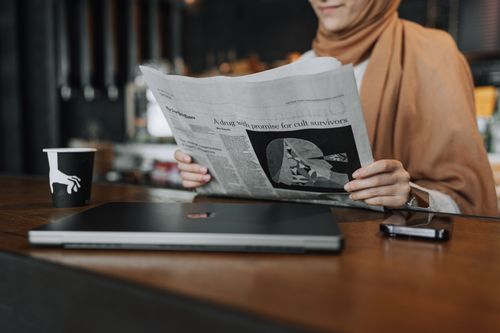Free A woman in a café reading a newspaper, with a coffee cup and laptop nearby, wearing a hijab. Stock Photo