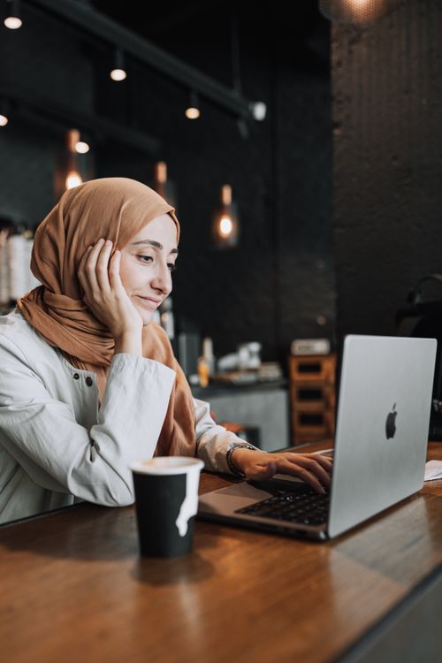 Free A woman in a headscarf works on her laptop in a warm café setting, enjoying a coffee. Stock Photo
