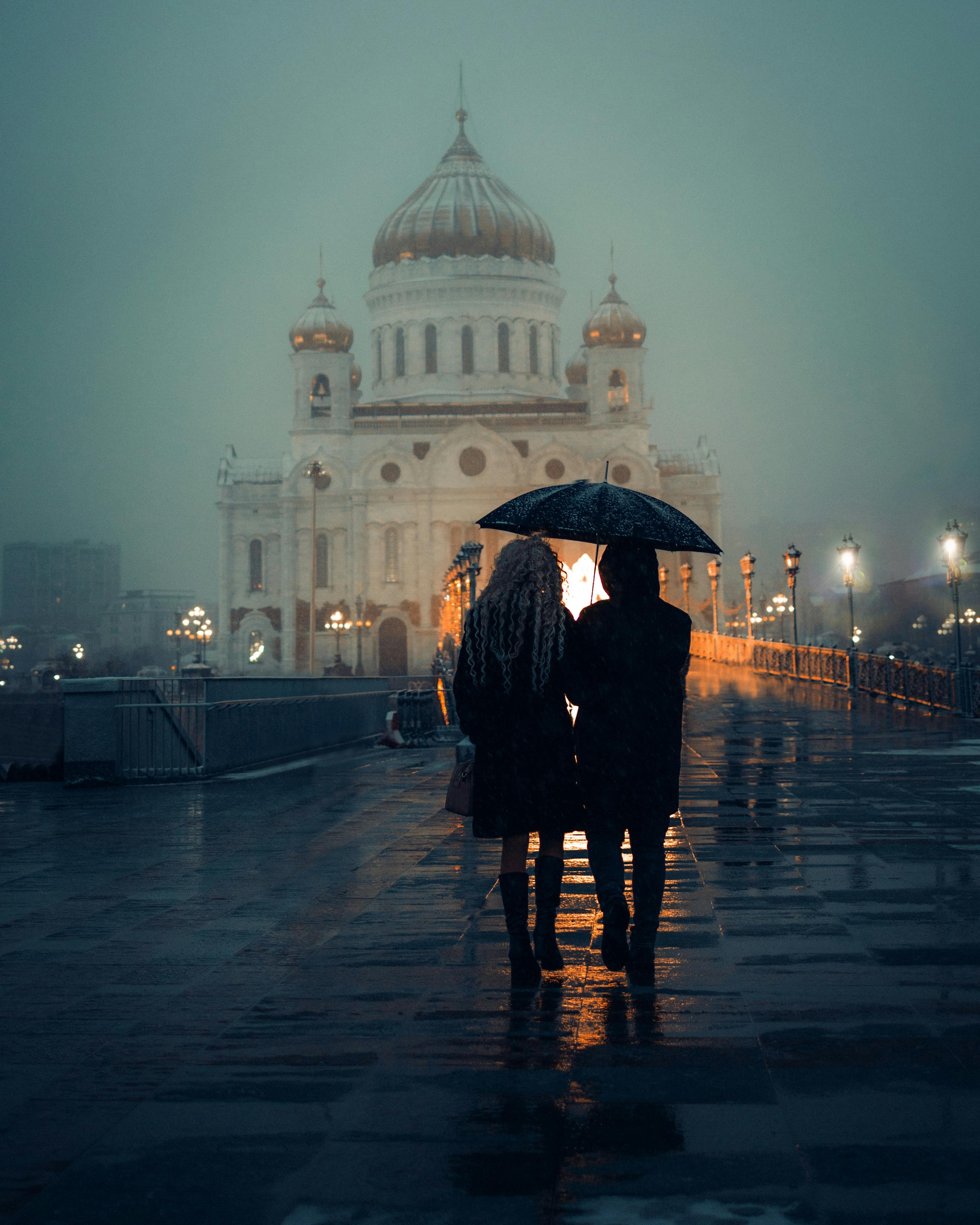 Couple under umbrella walks towards illuminated cathedral on a rainy evening.