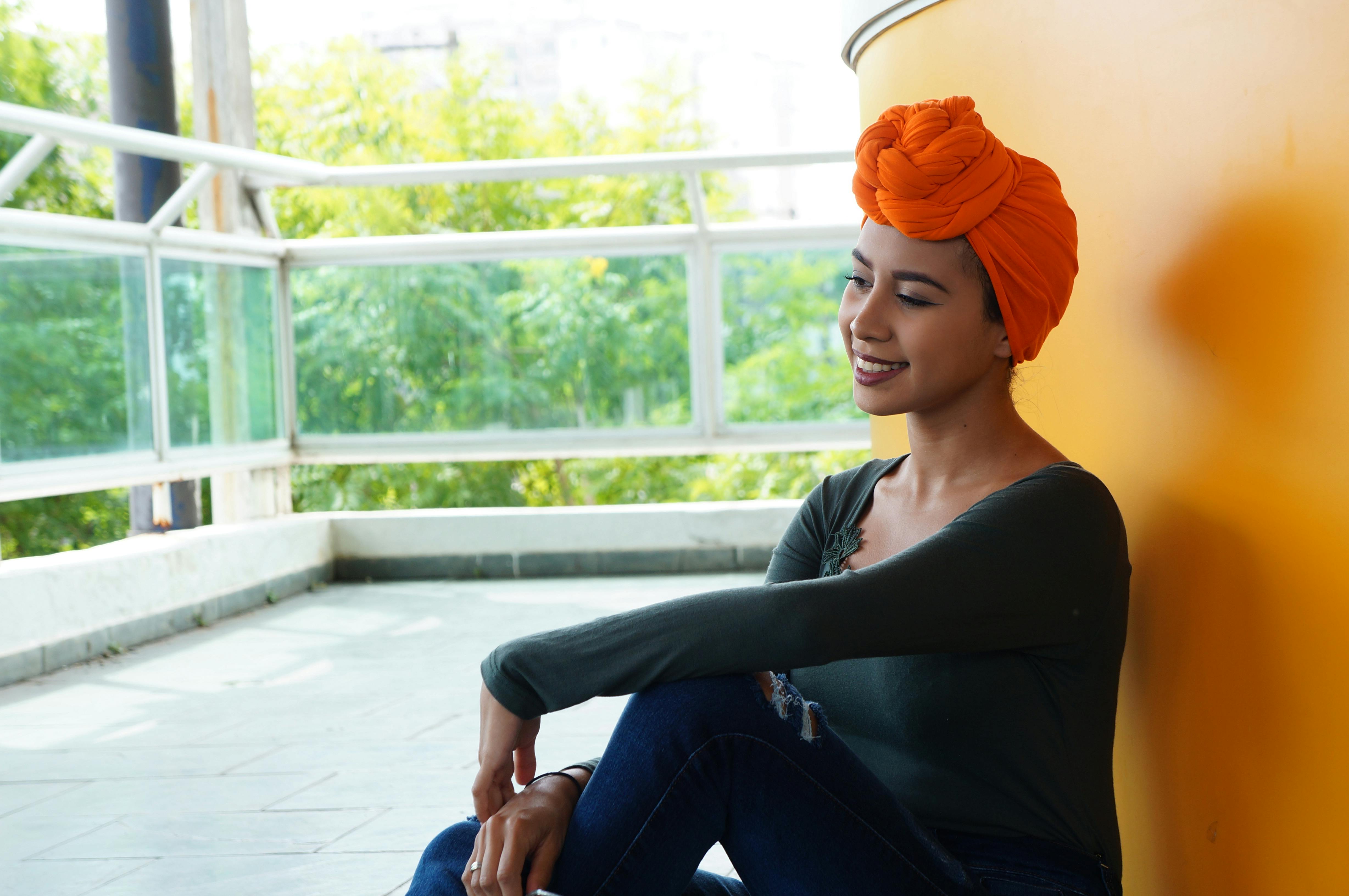 一位穿着休闲服装、戴着橙色头巾的女士面带微笑，坐在室内明亮的墙边