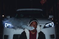 夜间，一名身穿连帽衫的男子在一辆开着车头灯的豪华车前吐着烟