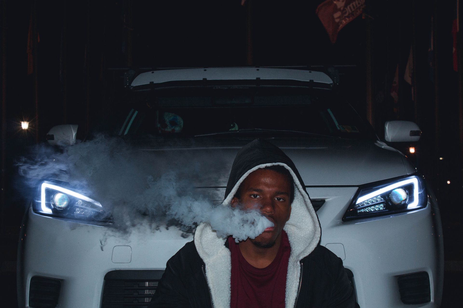 Man in hoodie exhaling smoke in front of luxury car with headlights at night.