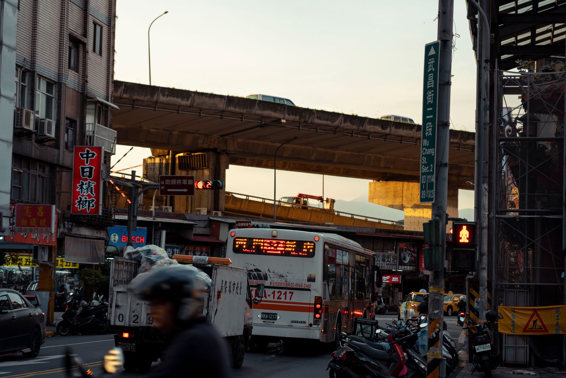A lively city street scene featuring traffic, including a bus, with an elevated road in the background.