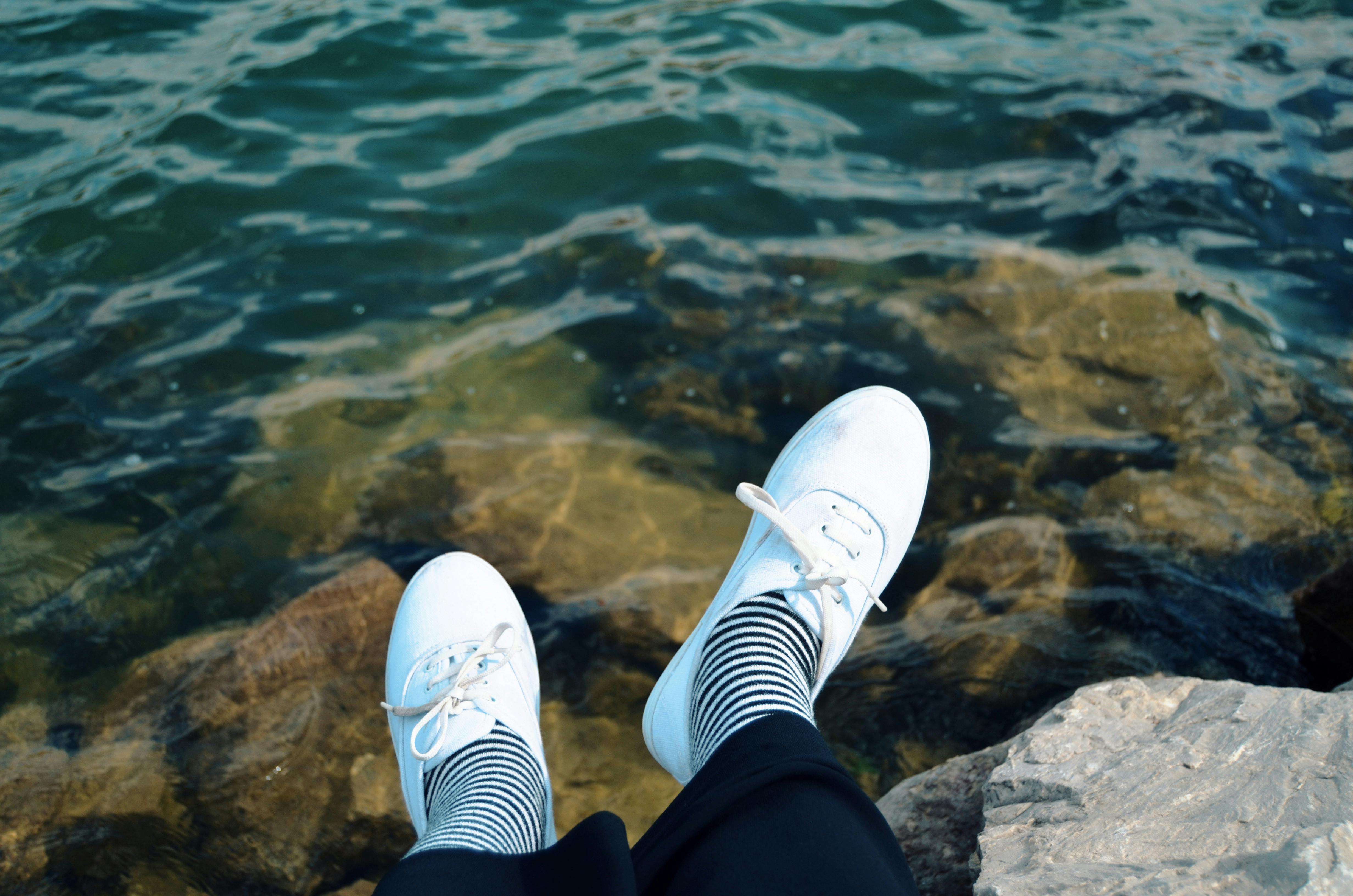Casual white sneakers paired with striped socks by the water on rocky shore.