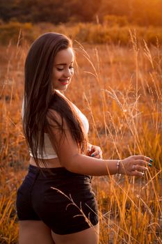 A woman in shorts smiles peacefully while standing in a sunlit field during sunset.