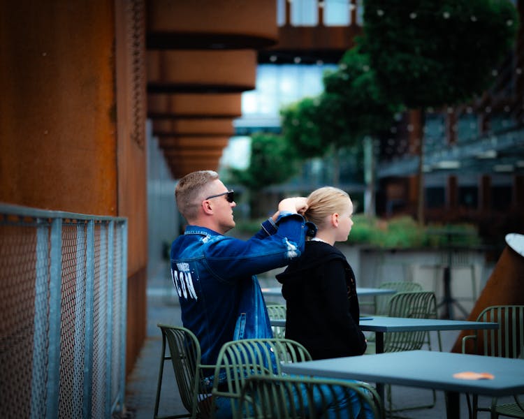 Father Tying His Daughters Hair While Sitting On A Chair In A Cafe Patio 