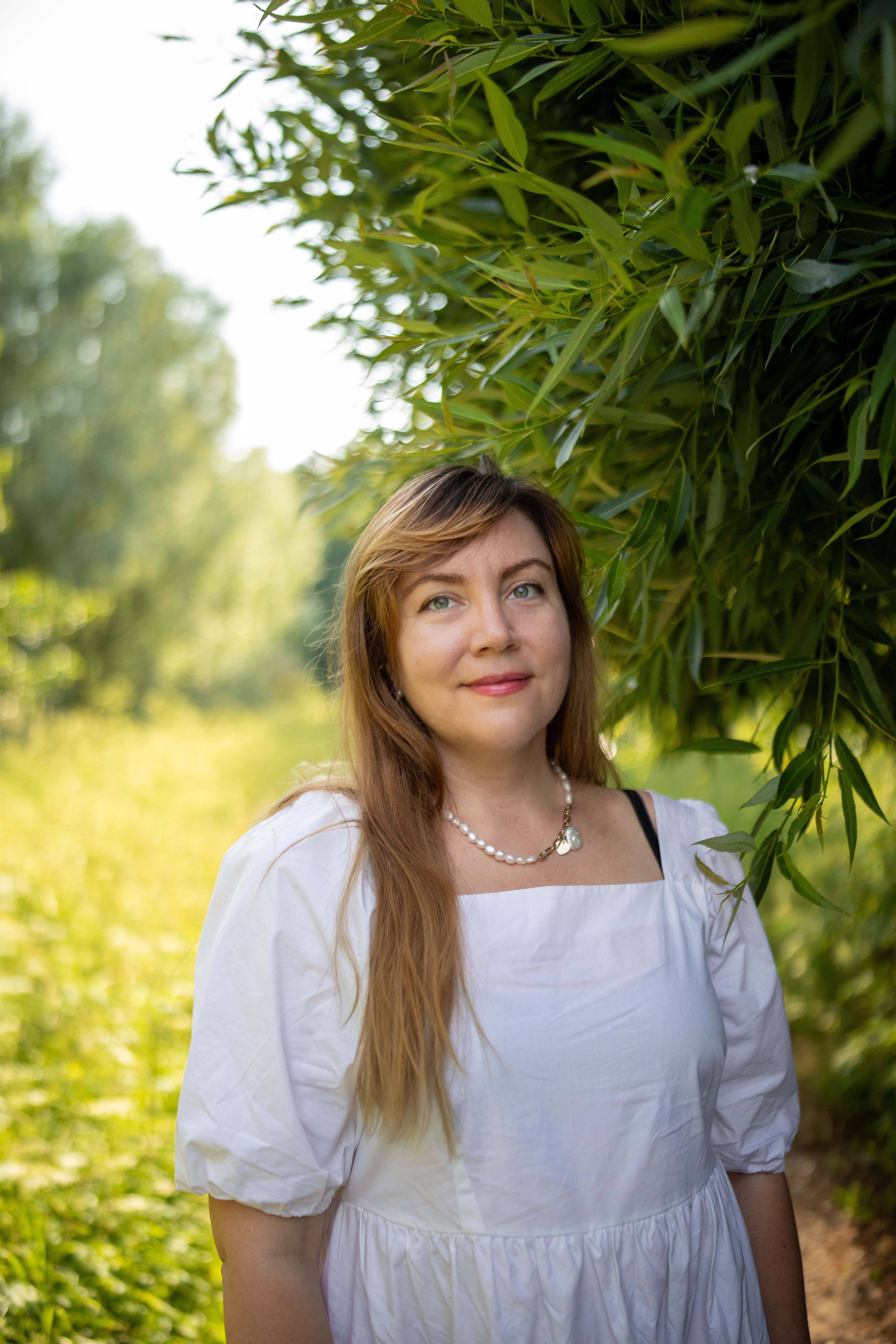 A smiling woman in a white dress standing in a sunny, green meadow.