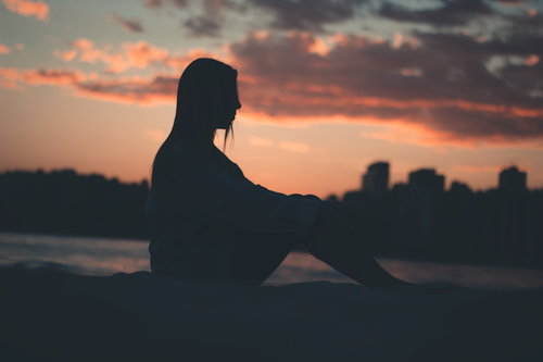 Free Silhouette of a woman sitting by the water at sunset with a city skyline in the background. Stock Photo