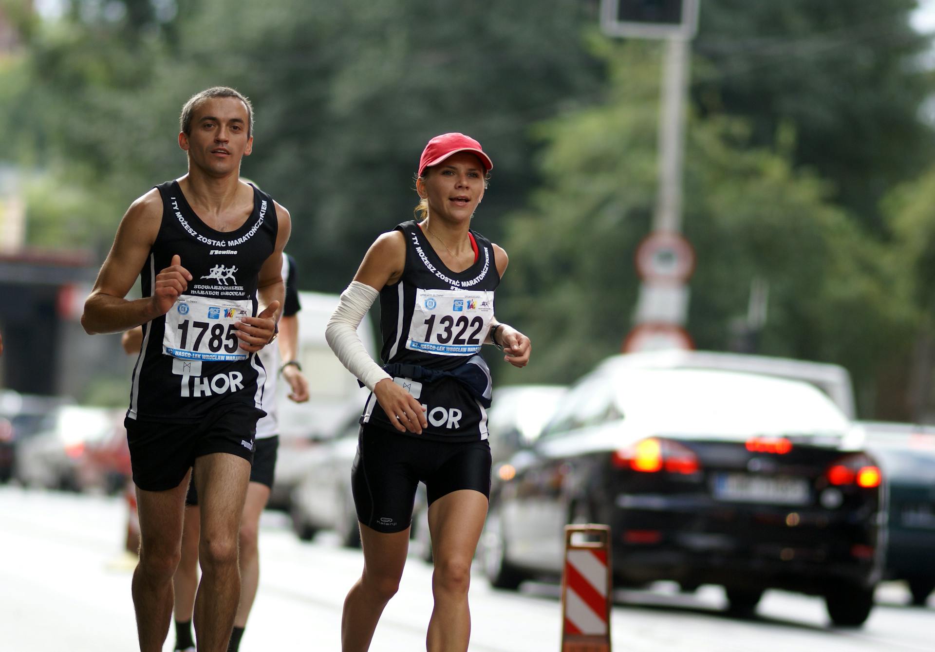 Two athletes running in a street marathon amidst urban traffic.