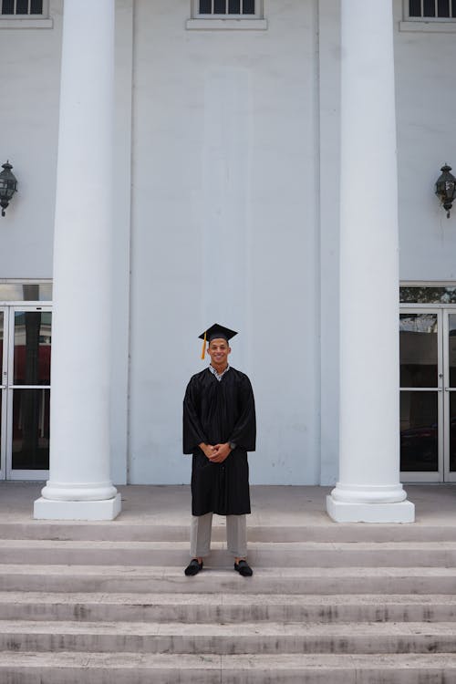 Young Man in a Gown and Mortarboard Standing on Steps in front of a Building and Smiling 