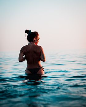 A woman stands topless in the ocean water at sunset, showcasing elegance and freedom.