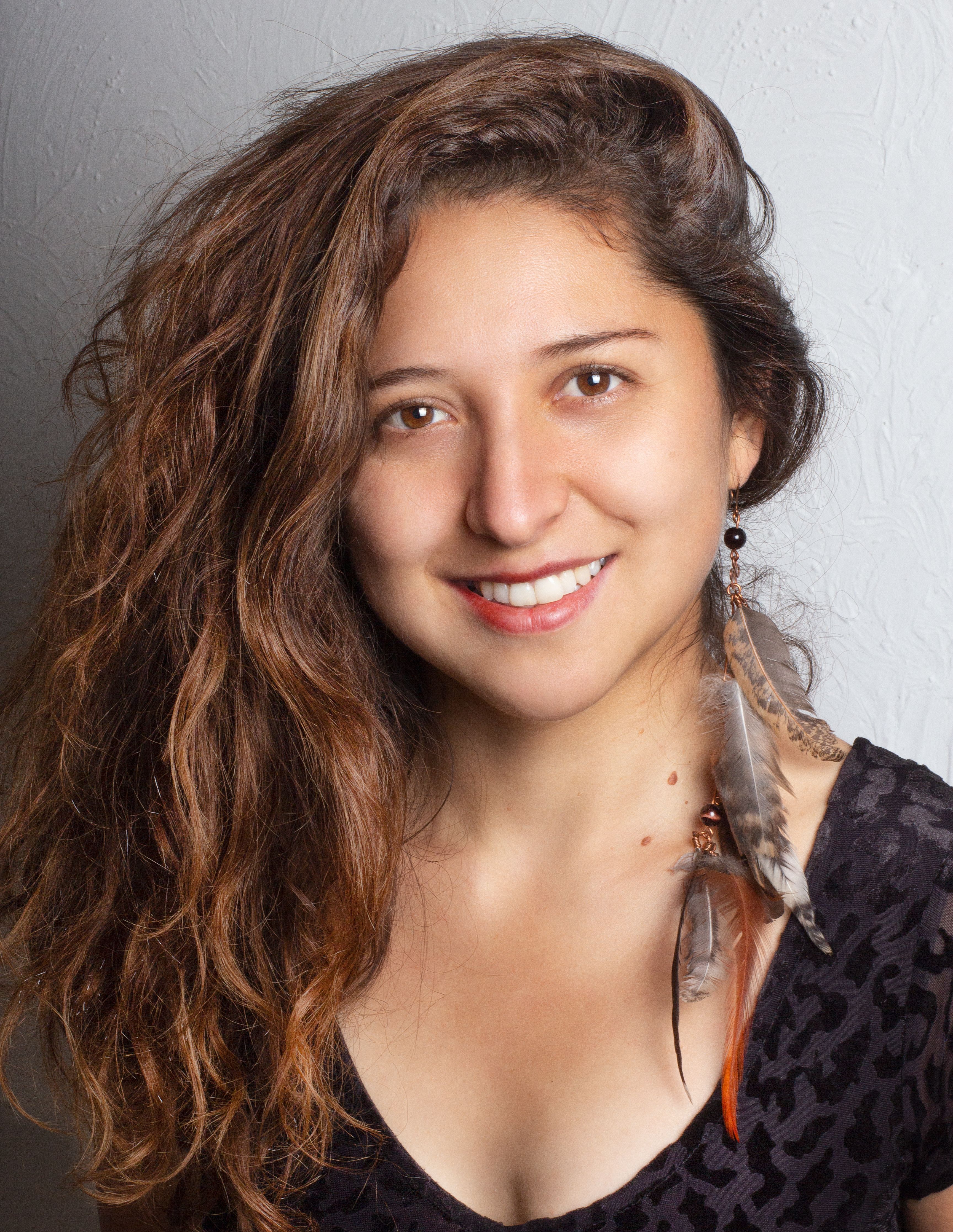 Young woman with brown hair and feather earrings smiling in a studio setting.