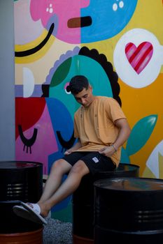 A young man in casual attire sits on barrels in front of a vibrant graffiti wall, urban scene.