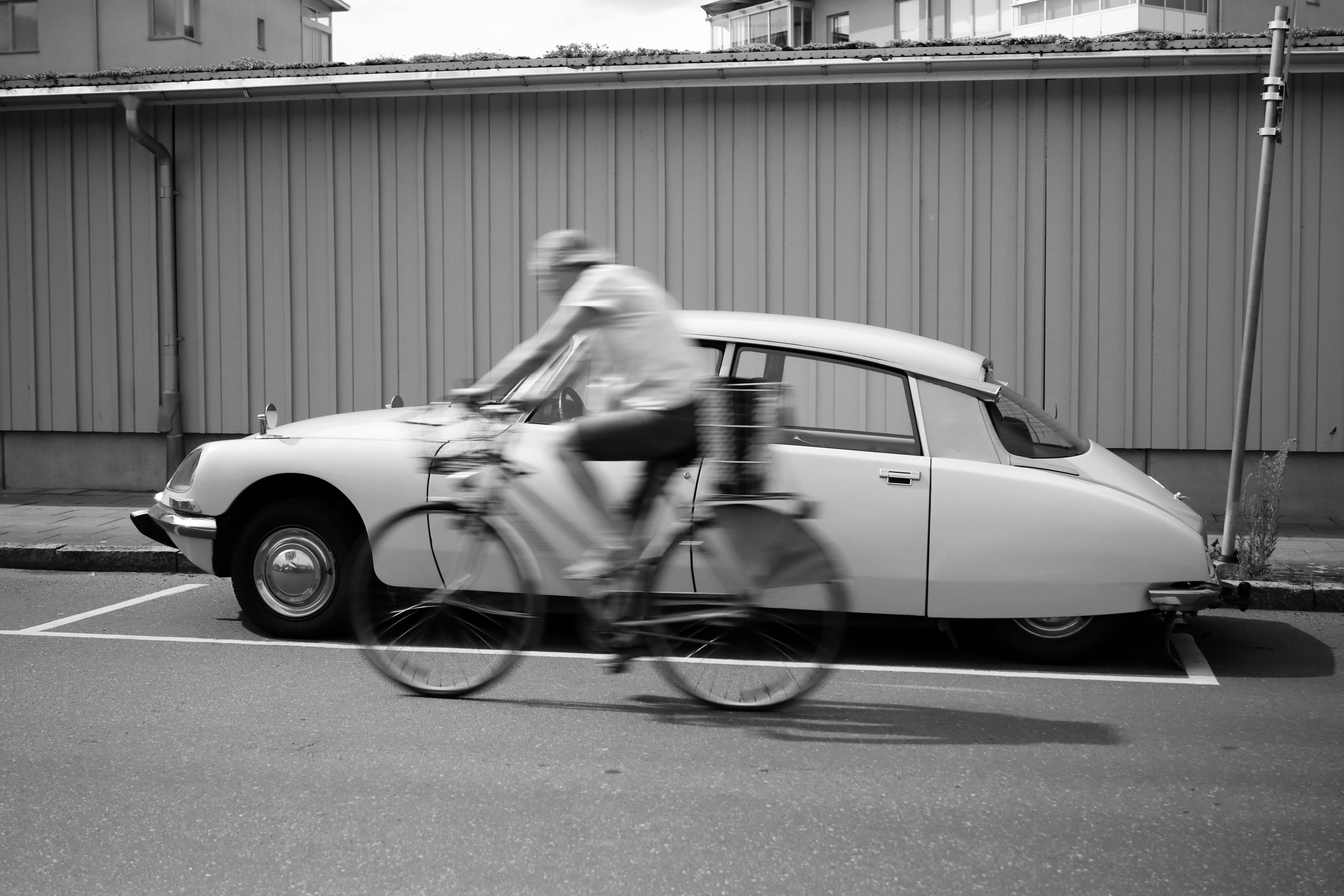 A vintage car parked on a city street with a cyclist in motion blur passing by.