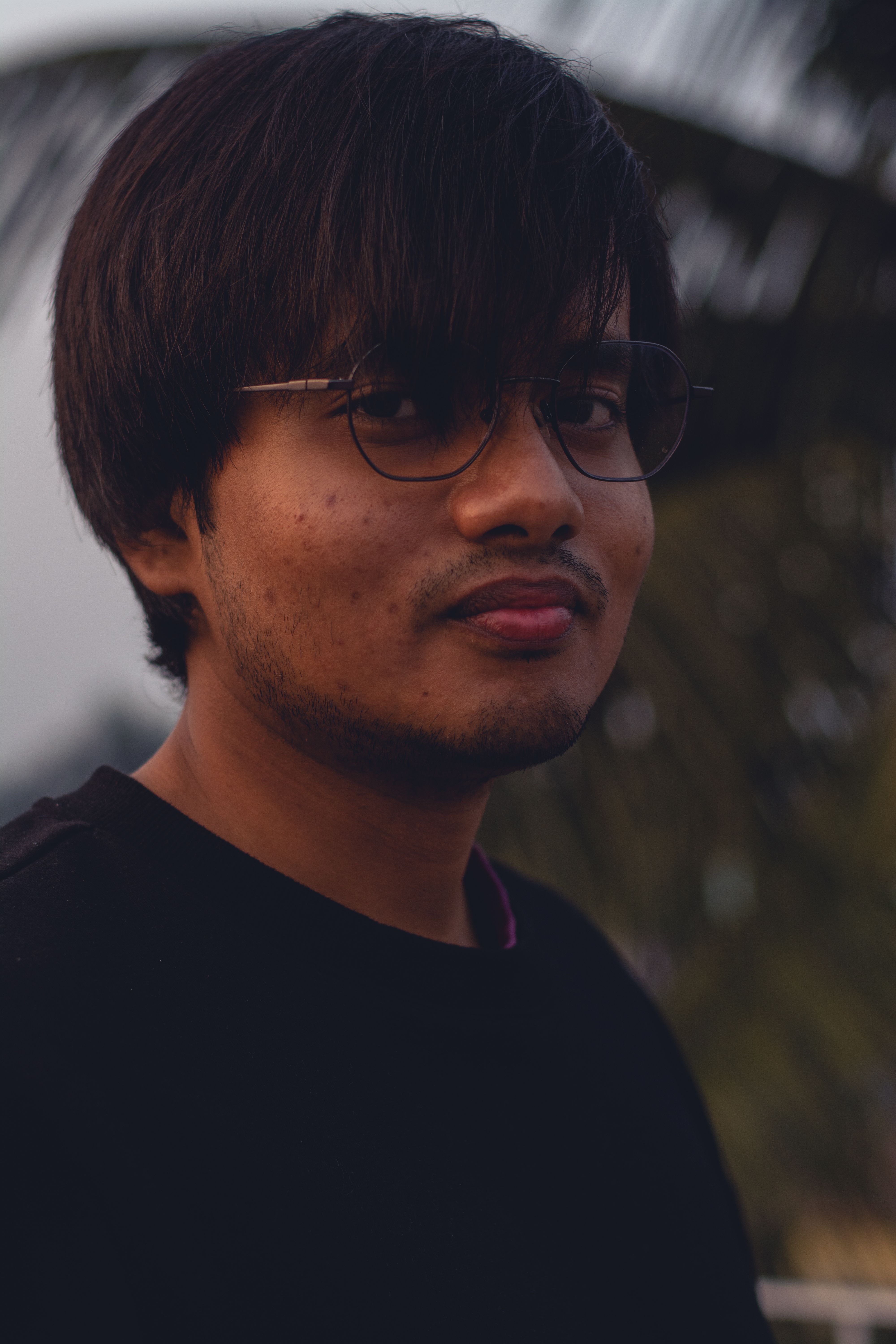 A portrait of a young man with eyeglasses standing outdoors at twilight, creating a moody atmosphere.